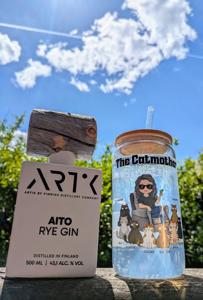 Strahlend blauer Himmel, ein paar Wölkchen und ein Kondensstreifen. Vor einer saftig grünen Hecke stehen eine Gin-Flasche (Aito Rye Gin 43,1% aus Finnland mit Holzdeckel, sieht ziemlich cool aus) und ein Trinkglas mit der Aufschrift "The Catmother". Auf dem Glas sitzt eine Comicfigur, die mir ähnelt, mit dunklen Locken, Sonnenbrille und einem Glas Gin Tonic in der Hand. Die Figur ist umgeben von acht Katzen: Mama, Elli, Pumba, Socke, Gauner, Shila, Milchbart und Pastete.