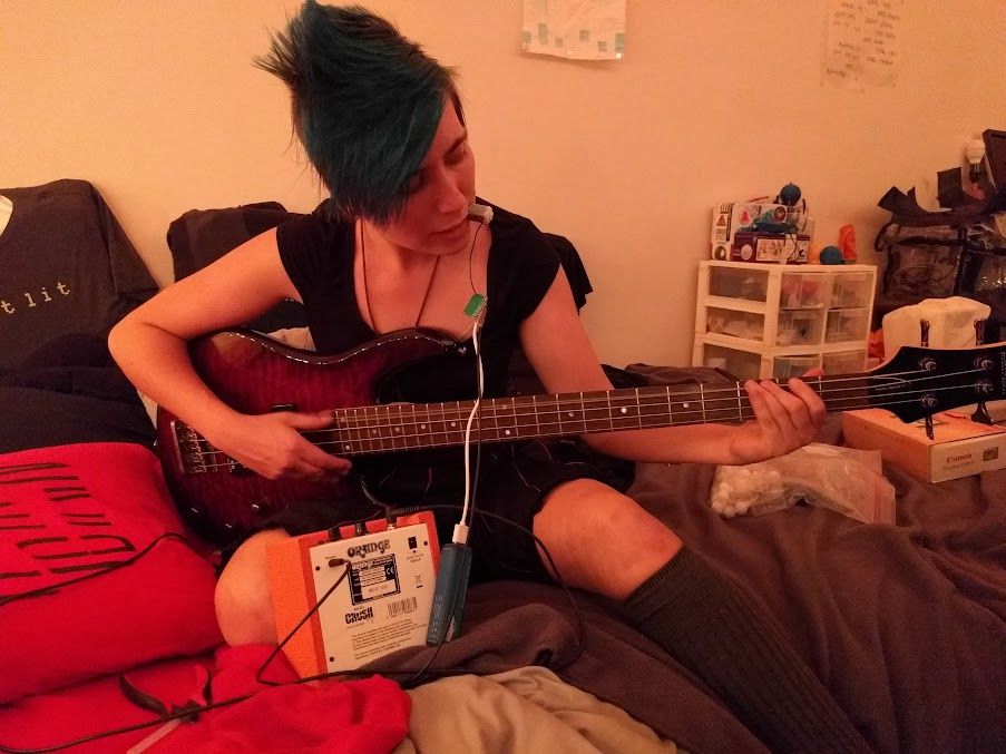 Alex, back when the hair stood up more, with a motor shaft clamped between her teeth, plucking a flame-red bass guitar that's plugged into a pathetically tiny Orange amp. A USB battery dangles from the motor, providing some extra Orange-flavored juice. Various art/tech projects clutter the area.
