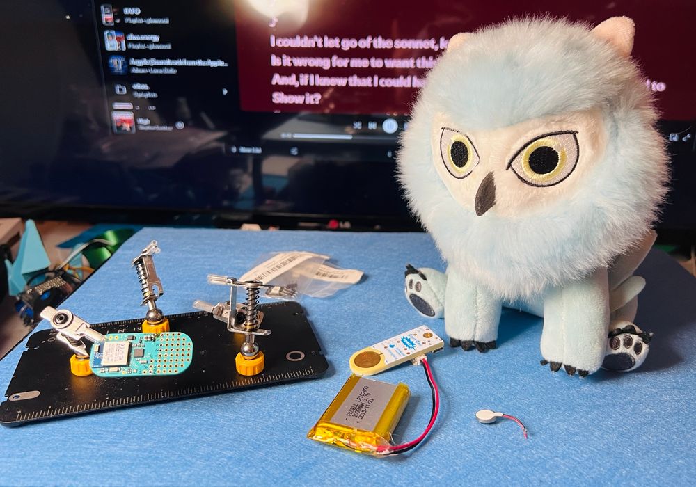 Desk with a LightBlue Bean on Omnifixo third hand, Coin Cell Power adapter + LiPo, haptic vibration motor, LEDs, and a stuffed light blue Owlbear! Lyrics to Stephen Sanchez's "High" in the bg.