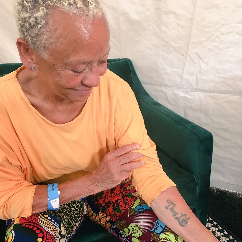 A Black woman with cropped hair and wearing an orange shirt displays a Thug Life tattoo on her forearm.