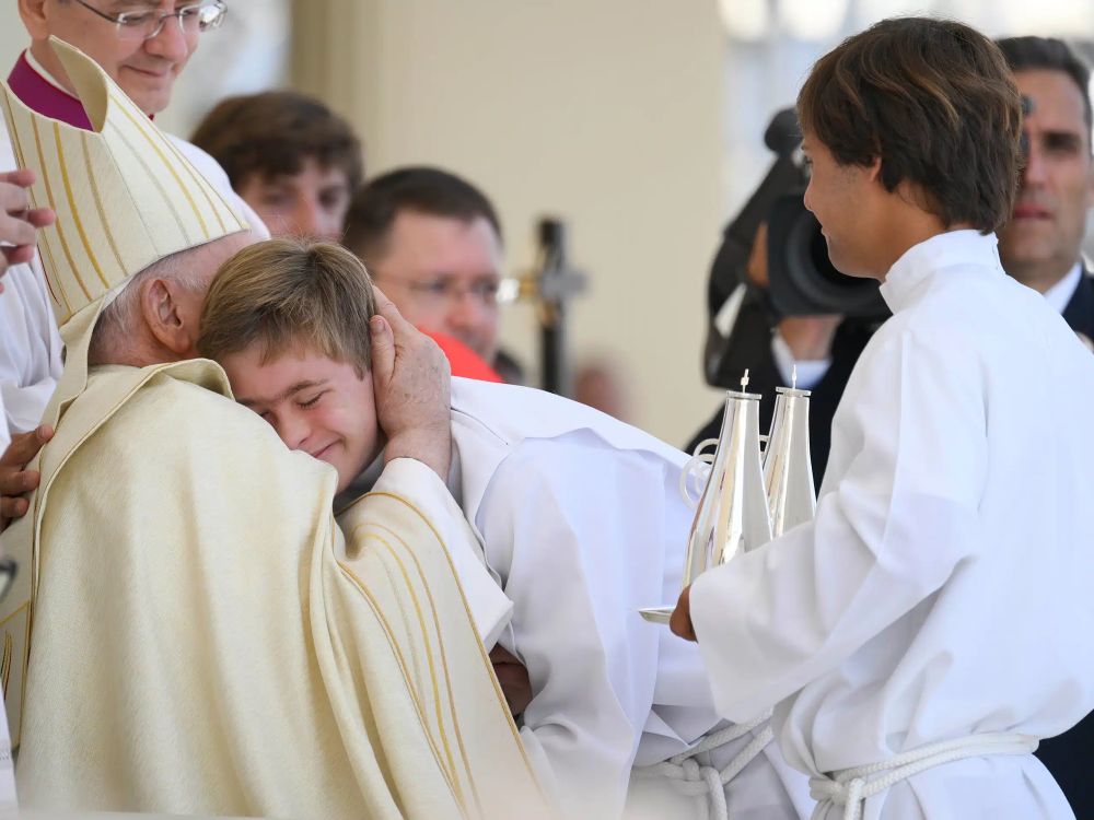 Pope embraces boy with Down syndrome. Boy has shaggy brown hair, his eyes are closed, but he is smiling. Pope’s face is covered by the boy’s.