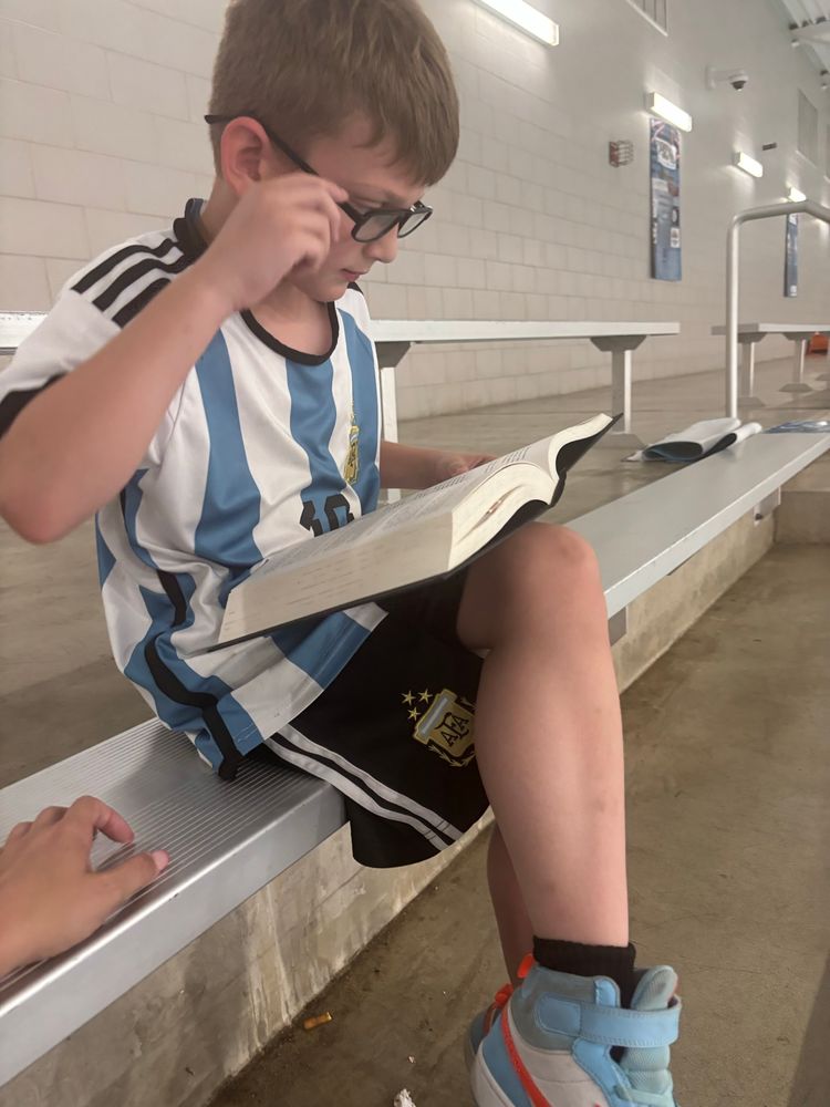 Blonde seven year old sitting on a bleacher with legs crossed and a large book on his lap and glasses on his face