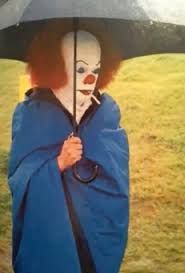 Tim Curry as Pennywise wrapped in a blanket under an umbrella and smoking a cigarette