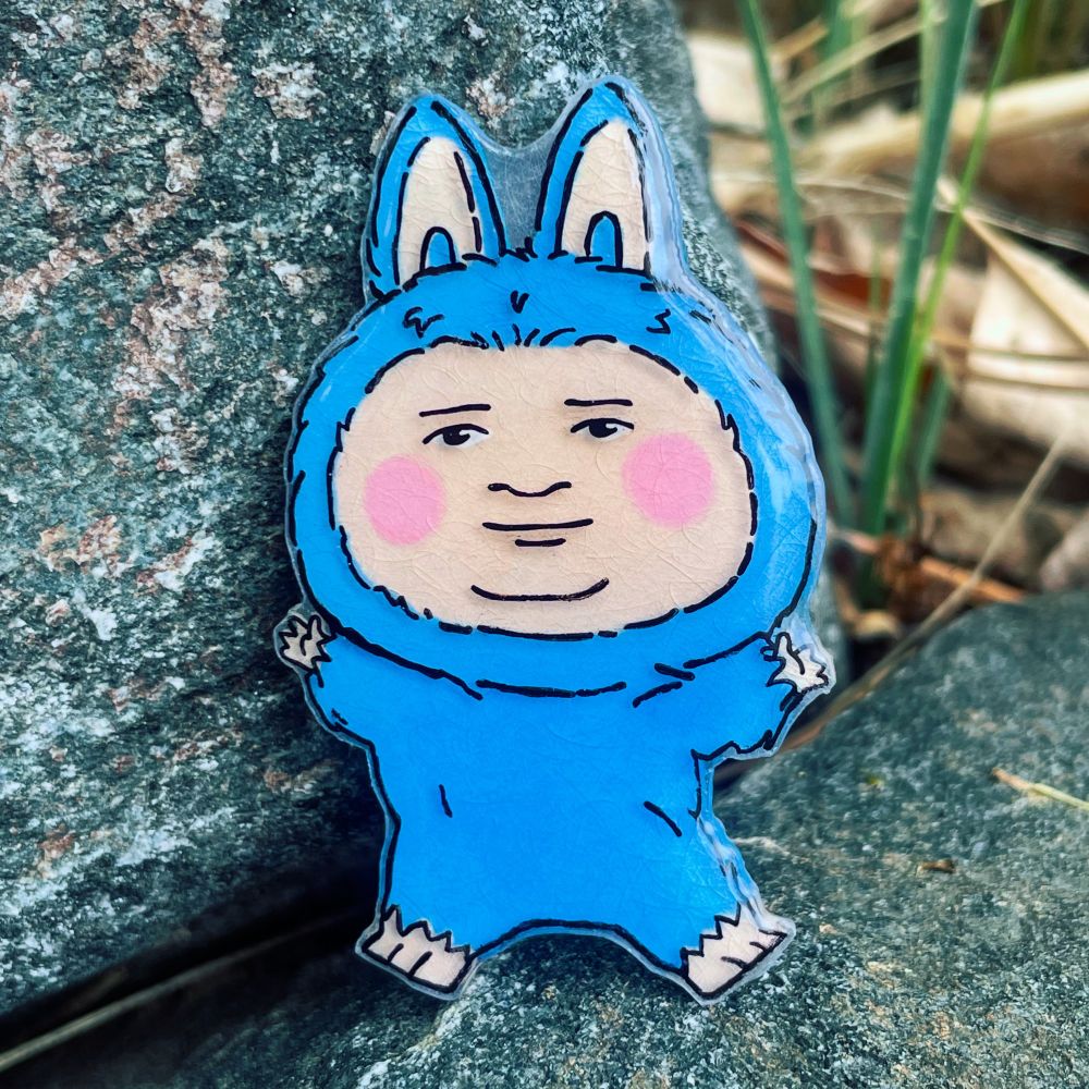 Bobby Hill’s Face is drawn on a blue Labubu’s body on shrink plastic and coated in UV resin. 