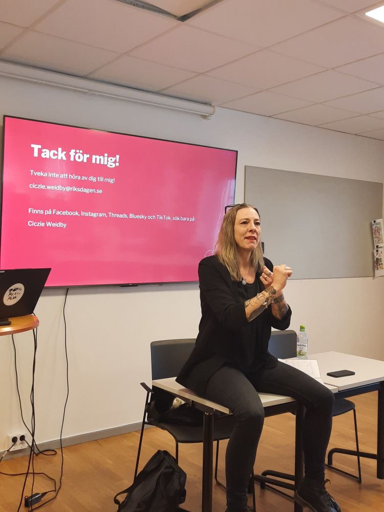 Ciczie sitter avslappnad på ett av konferensrummets bord, hon har de svartrimmade glasögonen i håret, och håller händerna framför sig som om hon räknar upp olika förslag. Bakom sig har hon den avslutande bilden i PowerPointpresentationen som det står "Tack för mig!" och hennes kontaktuppgifter samt sociala mediakonton på.