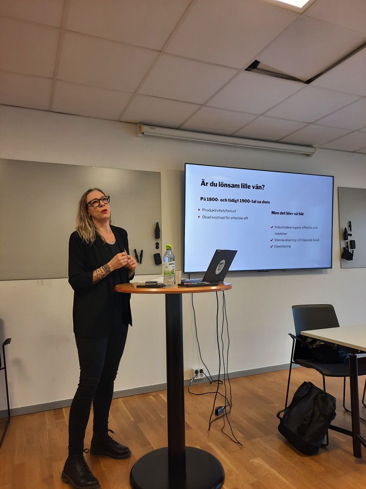 Ciczie, helt klädd i svart och med svarta glasögon, står och talar om arbetstidsförkortning. Hon står vid ett högt skrivbord med en laptop och vatten på. På skärmen bakom henne syns en slide från hennes PowerPointpresentation. På skärmen står det "Är du lönsam lille vän?" 