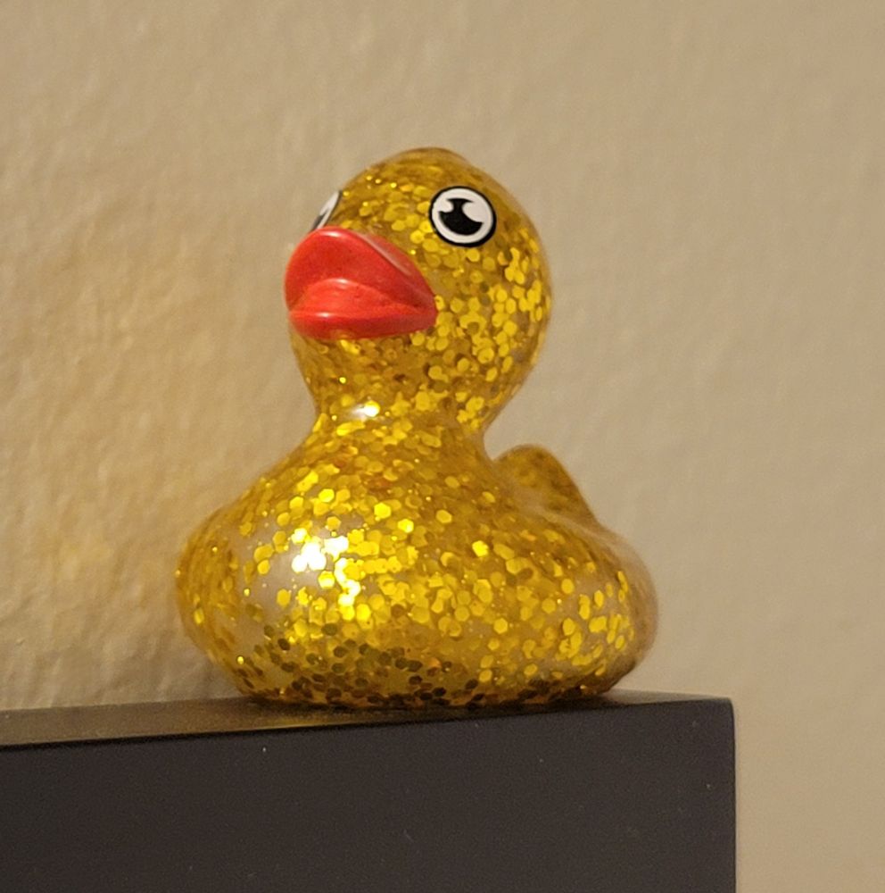 A full close up of the gold sparkly duck. The gold flecks look like small sequins and are embedded in the translucent rubber of the duck. The duck eyes look painted on and are very round. The duck is a bold contrasting orangey-red.