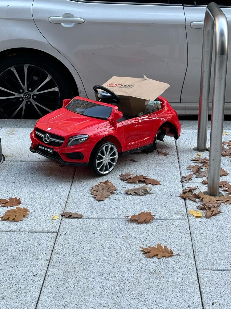 Photo of a red Mercedes toy car on sidewalk among leaves with bike rack to its right and a parked car behind it. Toy car is about the size of real car’s wheel.