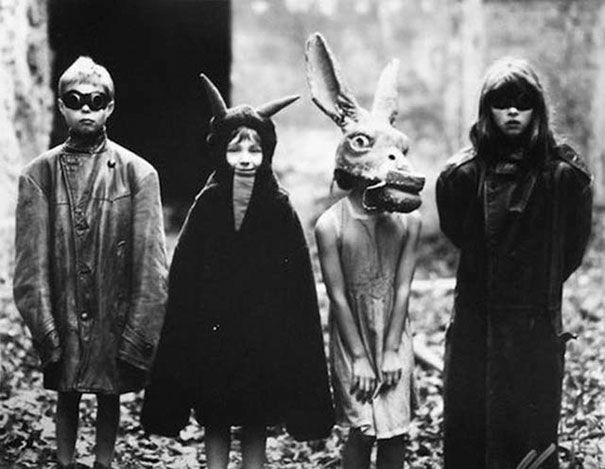 Four children in Halloween costumes. They are standing in a row. The two on each end both wear goggles and adult’s leather jackets. Another one is dressed as a devil, and the other one is a girl wearing a horse head mask. They are standing in a patch of waste ground between two buildings. I’m not sure where or when this photo was taken, although it’s probably American, late sixties or early seventies. It’s one of my favourite photos