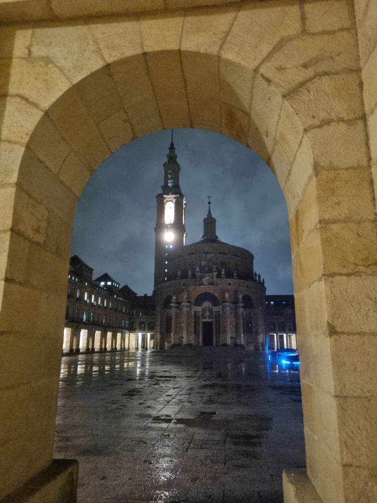 Foto de la Universidad Laboral tomada desde uno de sus pasillos. Se puede ver la torre del reloj iluminada y la iglesia, además del patio mojado. Al fondo a la derecha hay un coche con luces azules