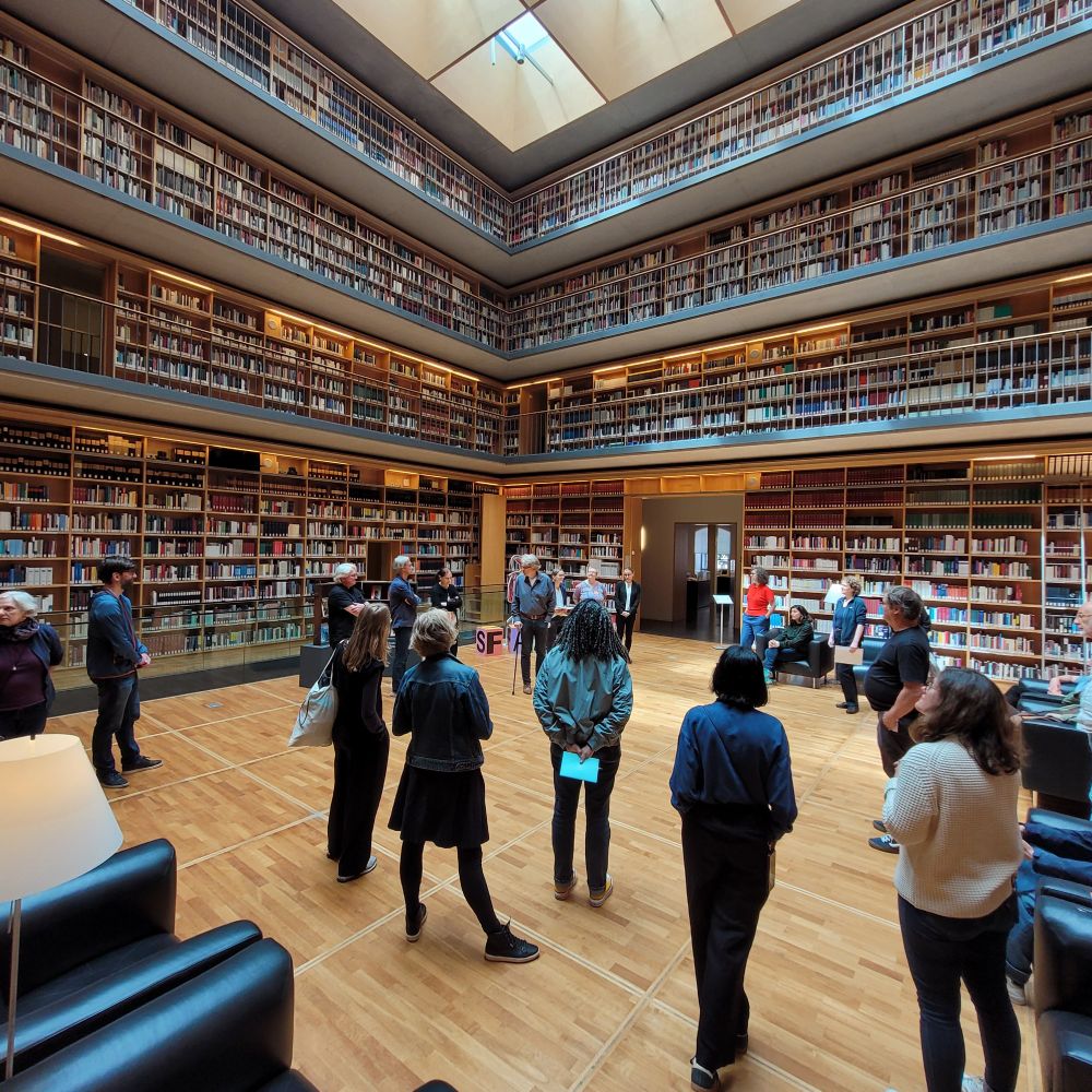 Großer quadratischer moderner Innenraum des Studienzentrums der Anna-Amalia-Bibliothek mit vier Galerie-Geschossen, die außen umlaufenden Regale stehen voller Bücher. Im Erdgeschoss begrüßen sieben Mitarbeiter*innen der Bibliothek unsere Kolleg*innen.