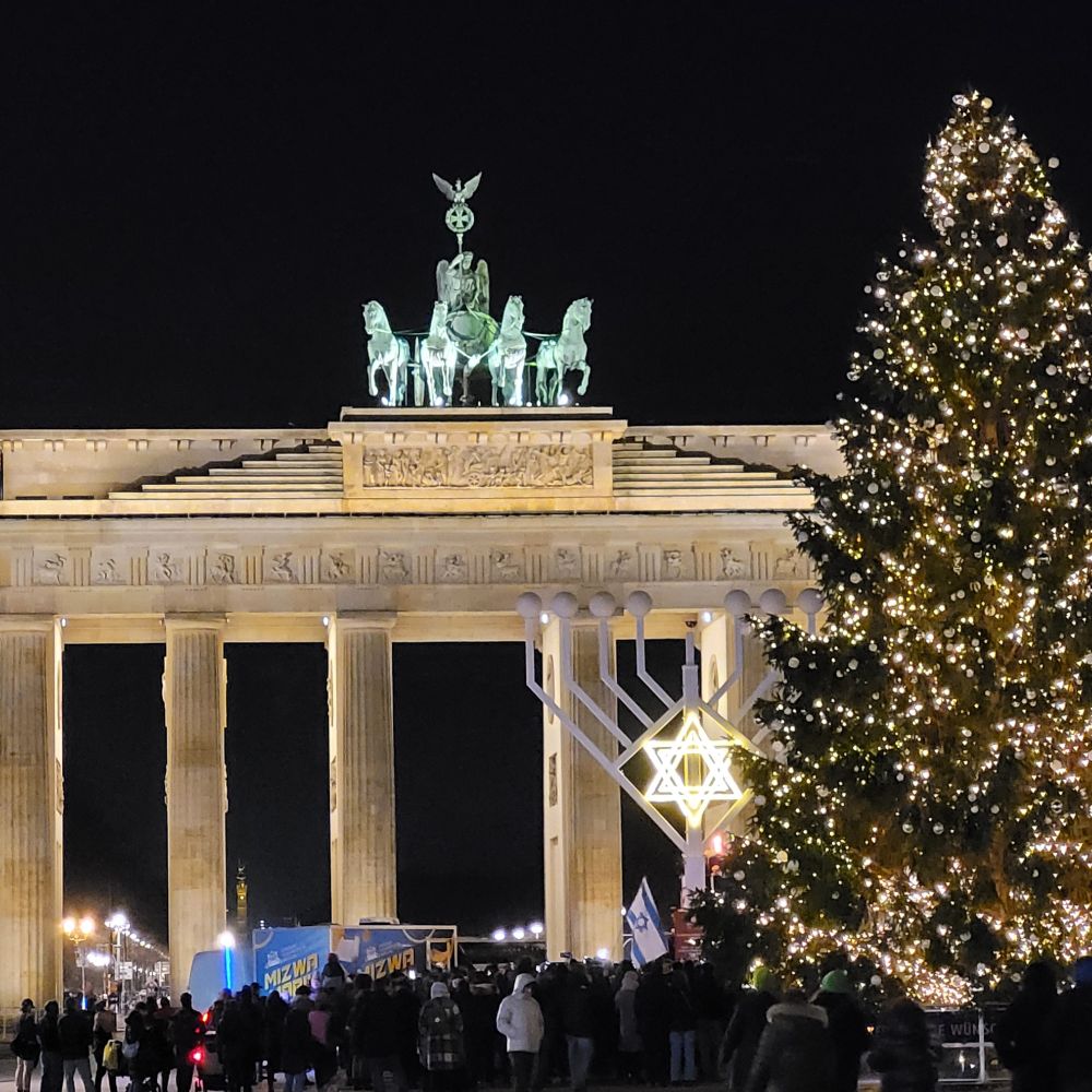 Im Dunklen im Hintergrund das Brandenburger Tor, davor viele Menschen, ein überdimensionaler Chanukkah-Leuchter mit beleuchtetem Davidstern und direkt daneben ein Weihnachtsbaum mit Lichterkette.