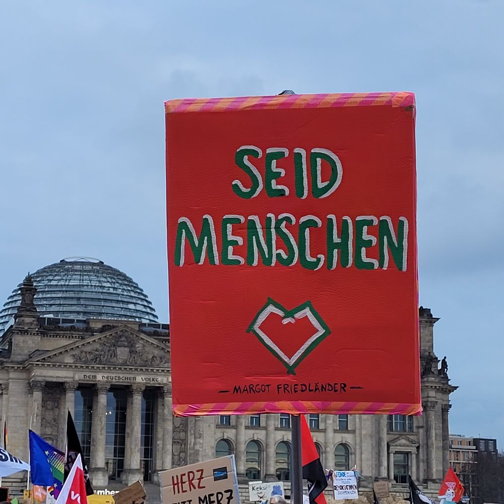 Reichstagsgebäude im Hintergrund, rotes Plakat, darauf in schwarz-weißer Schrift: Seid Menschen. Margot Friedländer