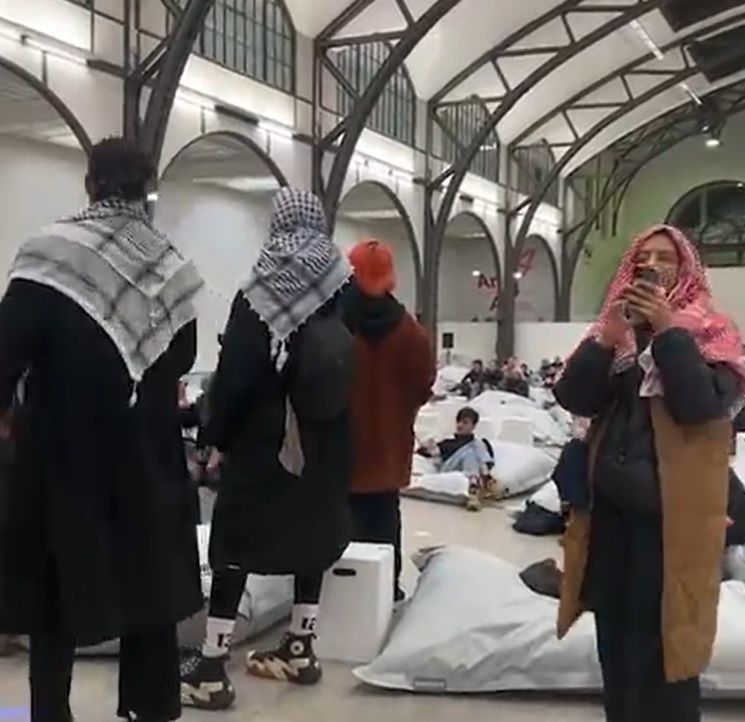 Publikum auf weißen Sitzsäcken im Museum Hamburger Bahnhof. Vor ihnen stehen vier Personen, drei tragen Palästinensertücher über Kopf und Schultern.