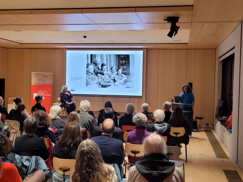 Rechts vor mehreren Reihen von Zuhörer*innen steht Autorin Ruth Preusse am Redepult und spricht in ein Handmikro. Links im Bild sitzt die Künstlerin und Mit-Autorin Inbar Chotzen. Auf der Großleinwand ist ein Schwarz-Weiß-Bild der Familie beim Kuchenessen zu sehen.