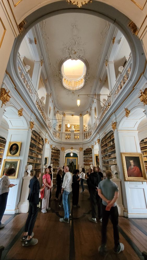 Blick in den barocken Bibliothekssaal der Anna-Amalia-Bibliothek: weiße Wände, goldene Bilderrahmen, goldene Regalbrett-Fronten, goldene Stuckelemente. Der Raum öffnet sich zu zwei darüber liegenden Galerien.