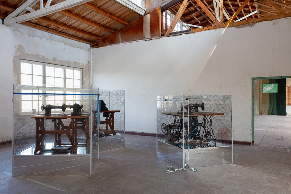 In einer Baracke mit Holzdach und großem Fenster im Hintergrund stehen drei Nähmaschinentische in drei gläsernen Vitrinen.