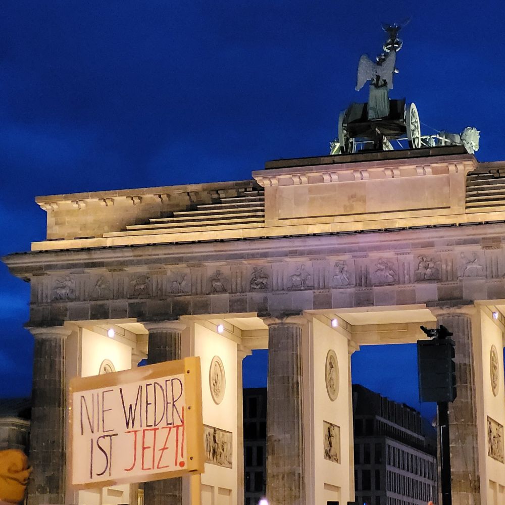 Angestrahltes Brandenburger Tor, davor Plakat mit Text Nie Wieder Ist Jetzt!