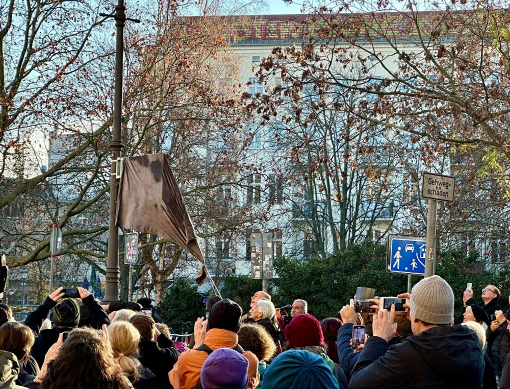 Eine Gruppe von circa dreißig Menschen beobachtet die Enthüllung eines Straßenschildes.
