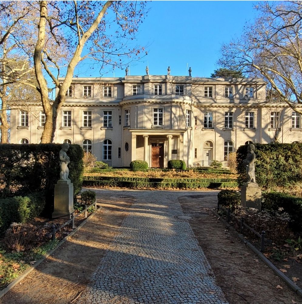 Sonnenbeschienene Fassade des Hauses der Wannsee-Konferenz, mittig der Haupteingang, blauer Himmel, kahle Bäume beiderseits des Weges zum Haus werfen Schatten auf die Fassade.