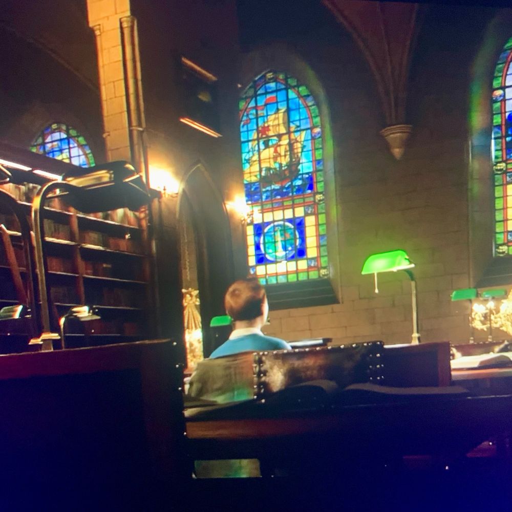 A still from The Adventures of Tintin (2011). Another angle of Tintin researching under the light of a chandelier in a cozy yet lavish library. You can see ornate wooden bookshelves and a stained glass window featuring designs of pirate ships. 
