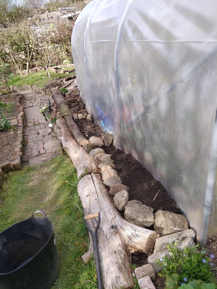 The side of a polytunnel in a garden. Rocks and logs have been placed along the edge to create flower beds
