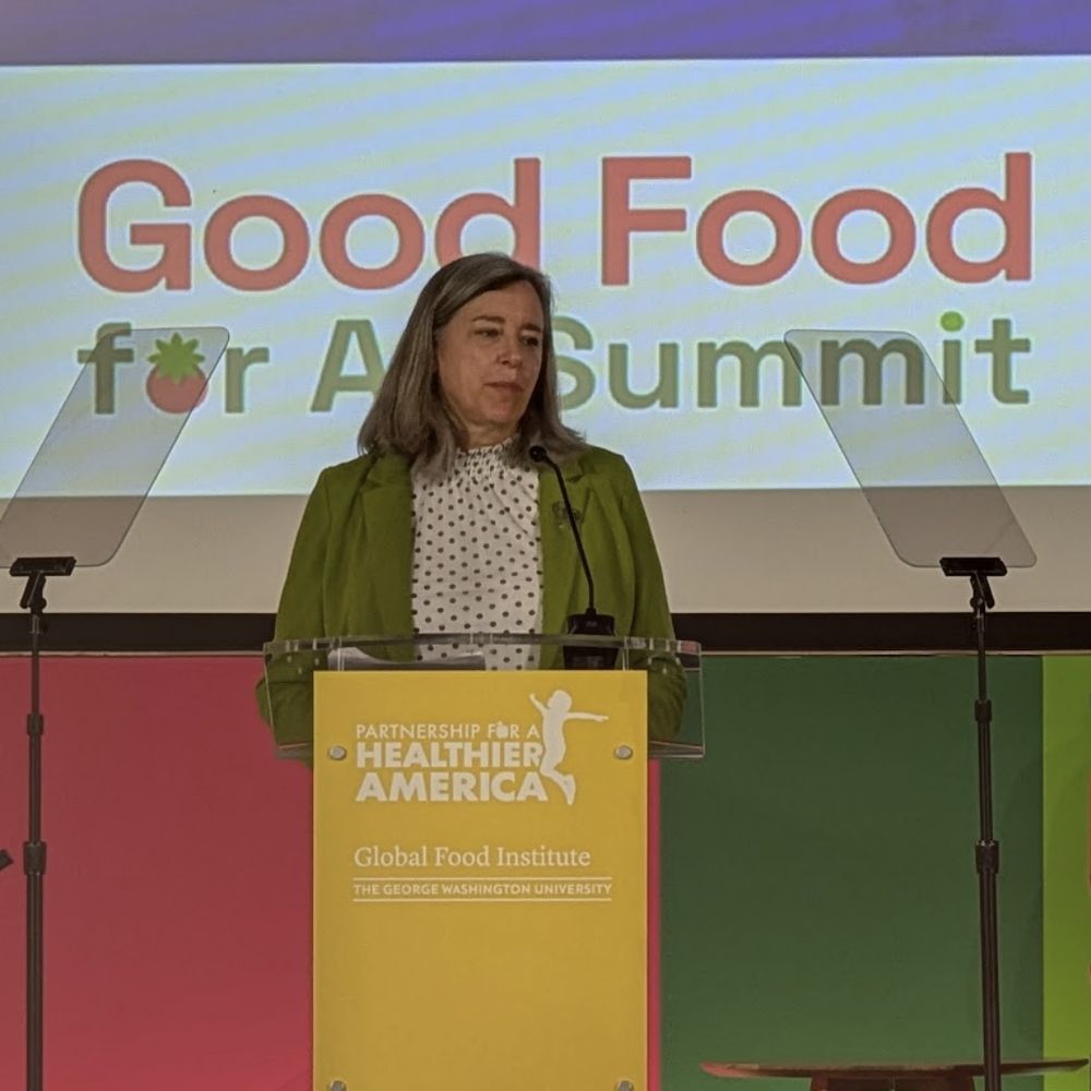 Stacy Dean of the Global Food Institute stands at the podium with the yellow sign, Partnership for a Healthier America. Behind her is a backdrop screen with the title, Good Food Summit 
