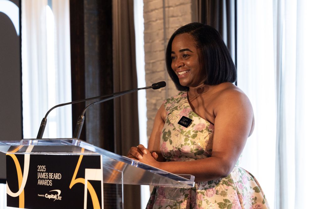 Seanicca Edwards Herron, a black American woman smiles at the podium. She is a wearing an off the shoulder, pastel floral printed evening gown. The clear podium has a black sign that reads, 2025 James Beard Awards sponsored by Capital One. The numbers 75 are in gold and yellow. Seanicaa, Founder and Executive Director of Freedmen Heirs Foundation Inc, received the James Beard Foundation's first Impact Award on June 16, 2025.