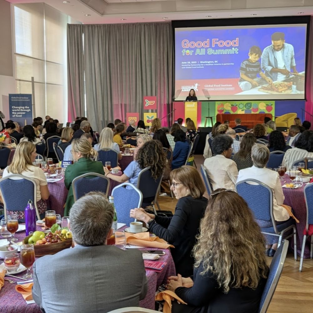 A conference room filled with people seated at round tables for the Good Food for All Summit 2025 hosted by Partnership for a Healthier America. They are looking at a speaker on stage with a backdrop image with a photo of a child and adult preparing food and the title, Good Food for All Summit.

