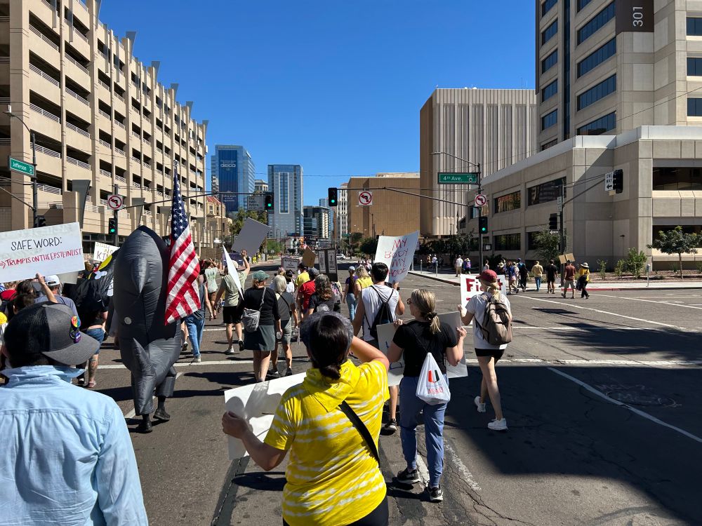 Protest march turning left at a stoplight. The protest includes a person in an inflatable shark costume holding an American flag, and many other people holding signs and marching.