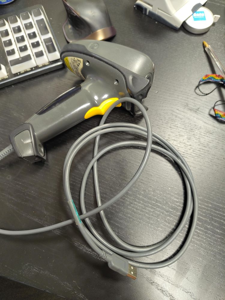 The Symbol/Zebra MT2070 barcode scanner lies on a black desk with its grey USB cable coiled around the handle, the yellow trigger visible.