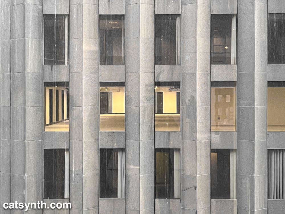 Looking at the side of a stone-clad office skyscraper.  Through the windows we see a completely empty office space with the lights on.