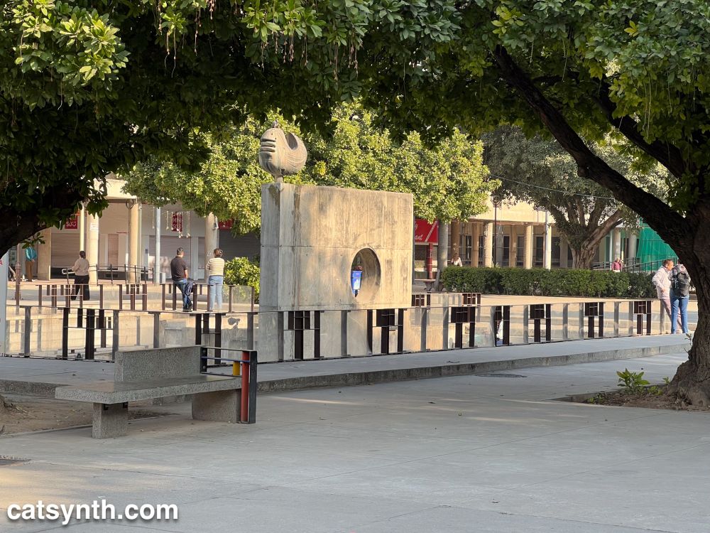 Concrete sculpture framed by trees in the middle of a concrete plaza with brutalist buildings visible in the background