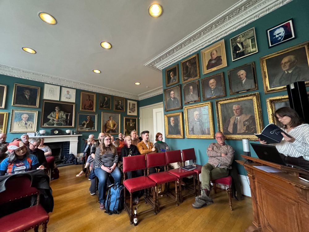 A room of audience members, Alison Irvine reading from a book, architect Malcolm Fraser, and many portraits on the wall.