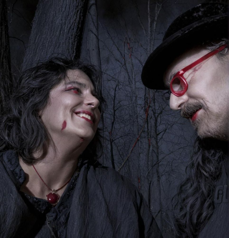 Kelli and David in cemetery with wooded background. Both have on black. David wears red glasses and a black bowler hat. Photo edited to simulate blood spatters.