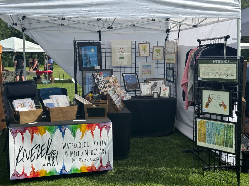 A booth full of colorful art, set up on the exceptionally plush grass of Ester Community Park, in Ester, Alaska