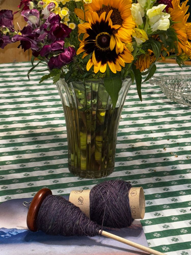 A spindle with a rich brown wooden whorl, half filled with purple yarn the rest of which has been wound off onto the cardboard core from a roll of toilet paper is resting on a green checked tablecloth in front of a vase of purple, yellow, and orange summer flowers.