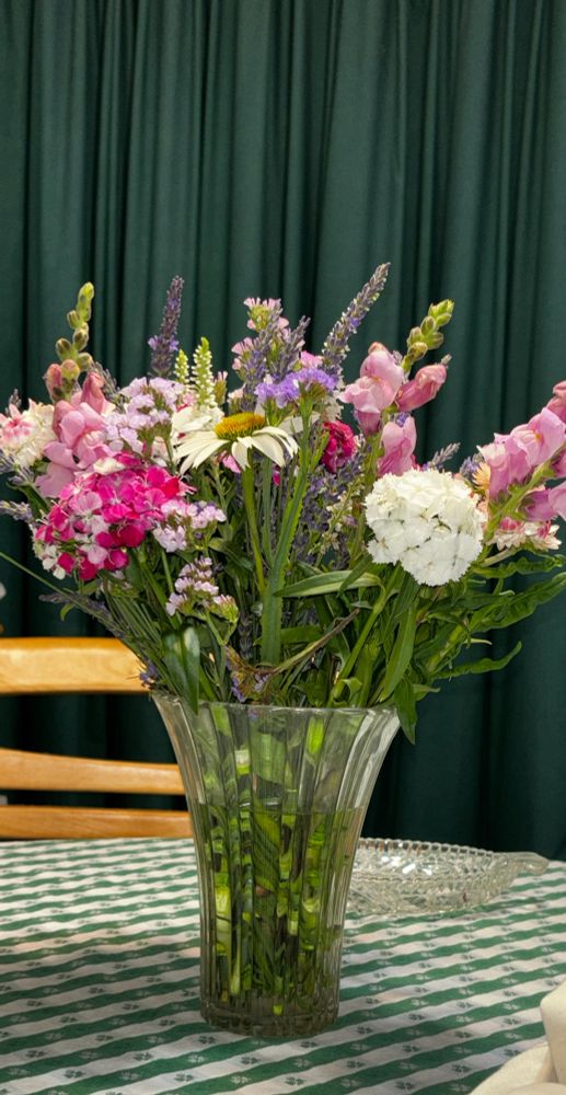 Pink, white, and purple flowers in a crystal vase, on a green checked tablecloth, with a dark green curtain in the background