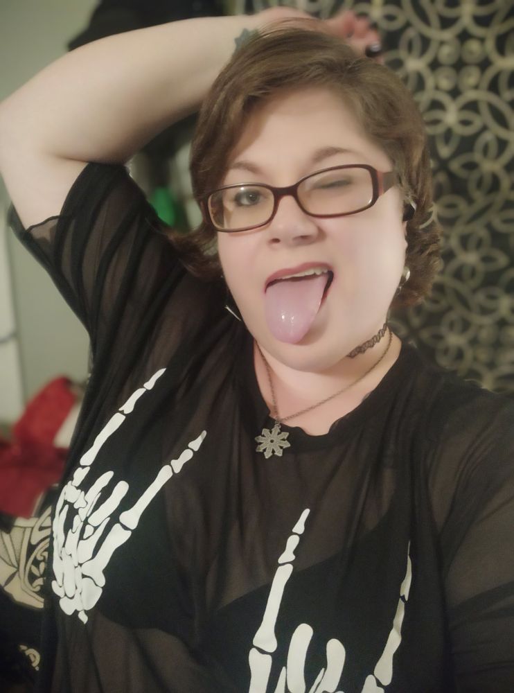 Vee poses for a silly portrait, with her right hand on her head, one eye closed, and her tongue sticking out. She's wearing a see-through black shirt with skeleton hands over the boobs that are making the "rock n roll" hand gesture.