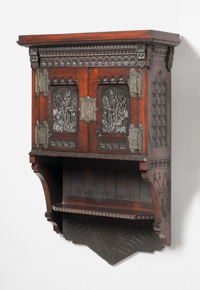 said wall cabinet's photograph. It has a lovely red wood and silver metal hinges. The inside of the door paneling appears to also have inner embossed silver panels inlaid into it. The various carvings and embossing have an organic, plant-based theme, with leaves and only a few flowers.