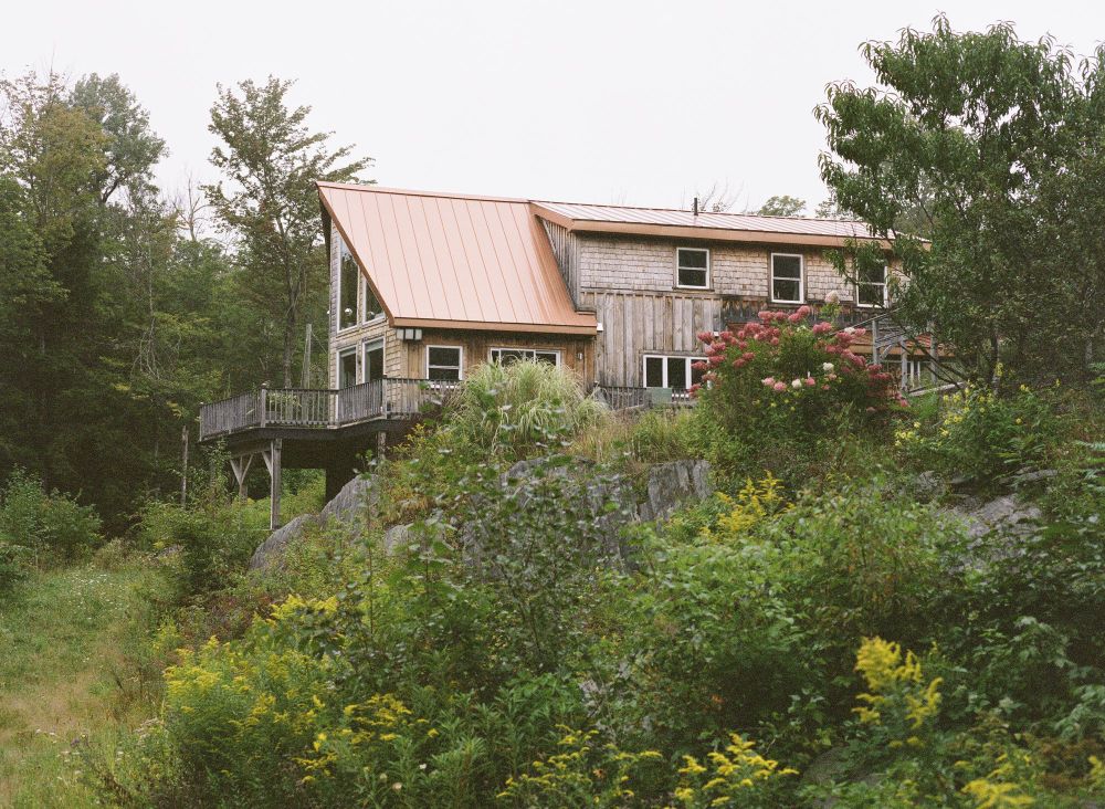 A picture of a large house viewable through early fall foliage. It has an orange metal roof, natural wood siding, wrap around deck and huge a frame style windows on one side. 
