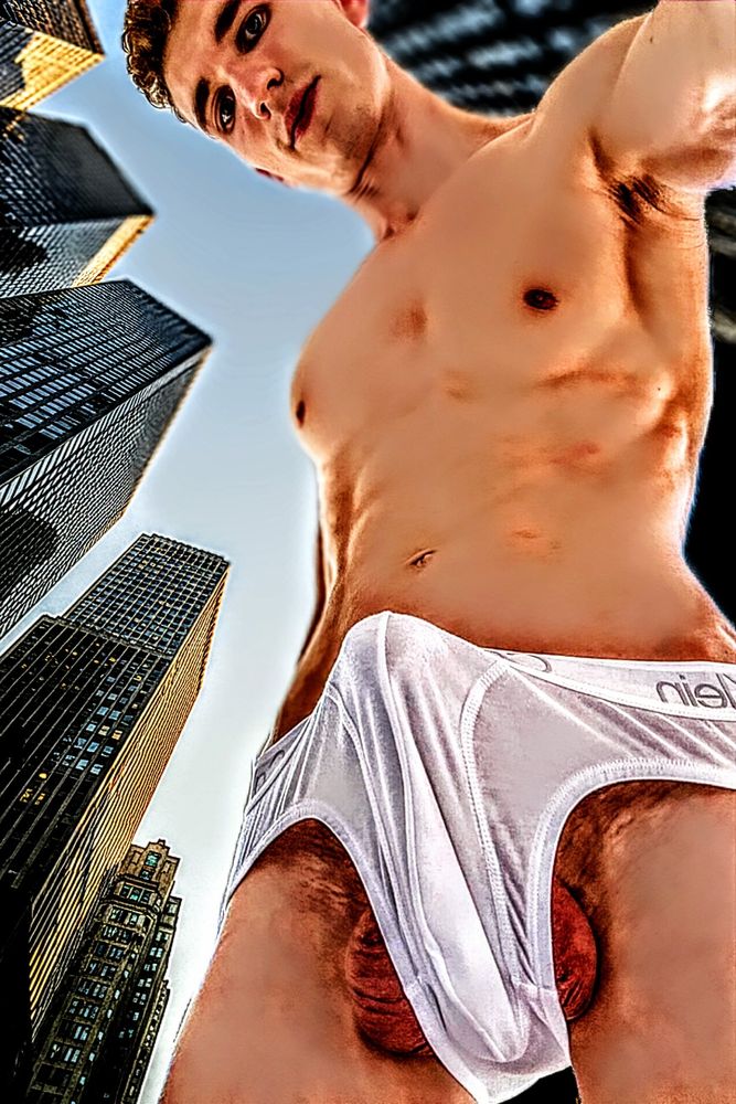 Upshot view of a male giant young college male model named lance, wearing only white Calvin Klein underwear that is bulging outward with his huge boner. His balls are hanging out and the underwear fabric is stretched to it's limits. Skyscrapers look above him in this upshot skyward angle while he looks like a giant watching others stare at his growing bulge from below.