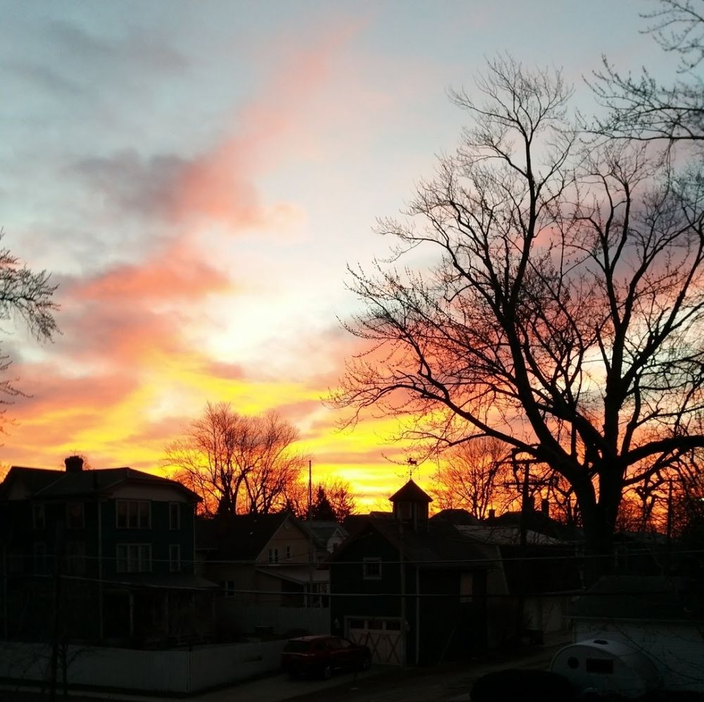 Rich yellow and orange hues stretching through a cloudy blue sky at sunrise over a heavily shadowed neighborhood of homes and trees
