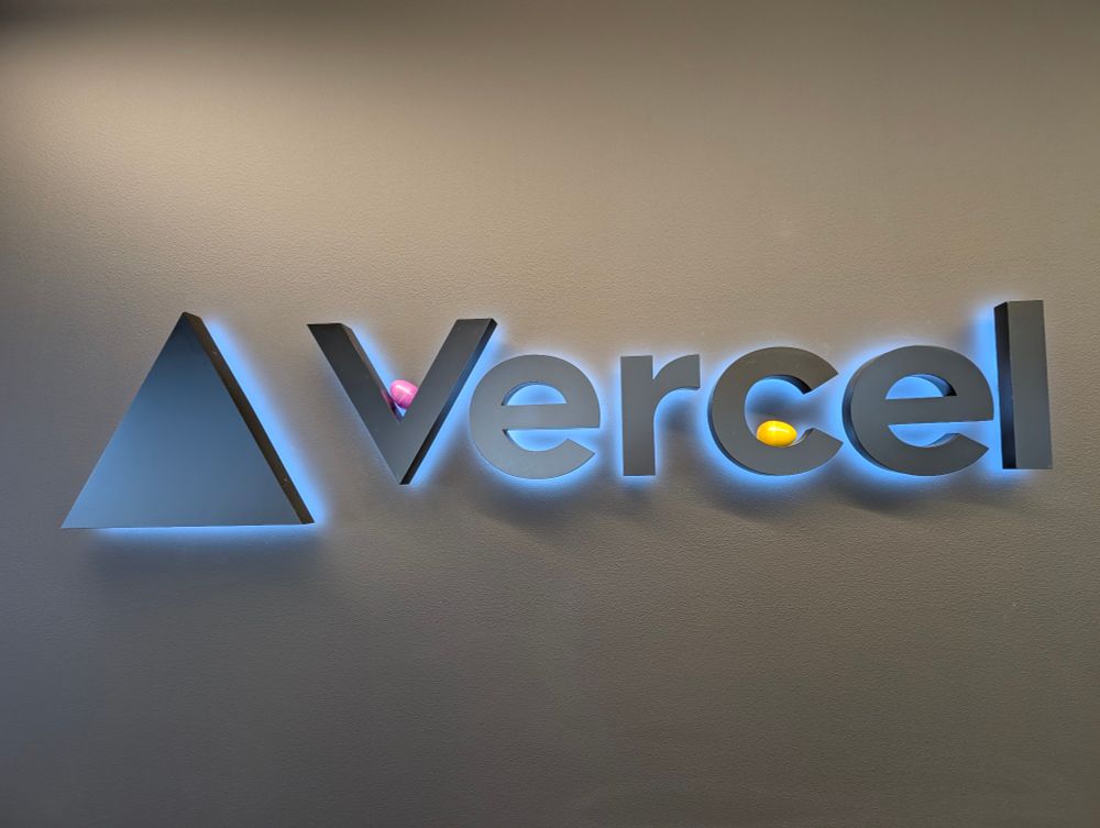 A photograph of a sign representing the Vercel logo and wordmark. There are two plastic Easter eggs: one pink one resting at the bottom of the 'V' and one yellow one resting at the bottom of the letter 'c'.