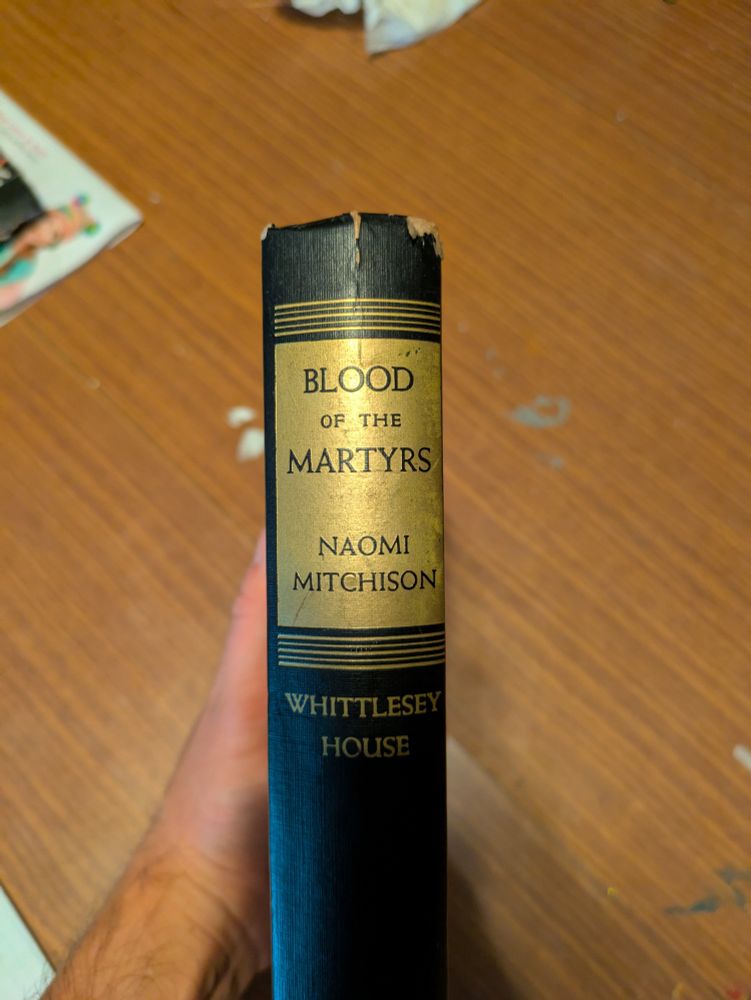 The spine of a black hardcover book. There are gilded stripes above and below a gilded rectangle with the title and author in black text. It reads Blood of the Martyrs Naomi Mitchison.