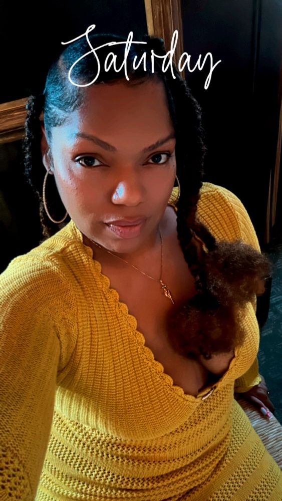 Black woman in a yellow knit dress, with large hoop earrings, a gold chain, and plaited ponytails with Afro puffs on the end 