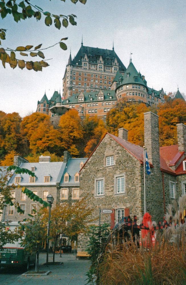 Une vue de château Frontenac dans la ville de Québec. La photo est prise depuis le bas de la vieille ville, on voit donc le château en contre plongée. Les arbres sont tout oranges, on comprend que l'on est en automne.