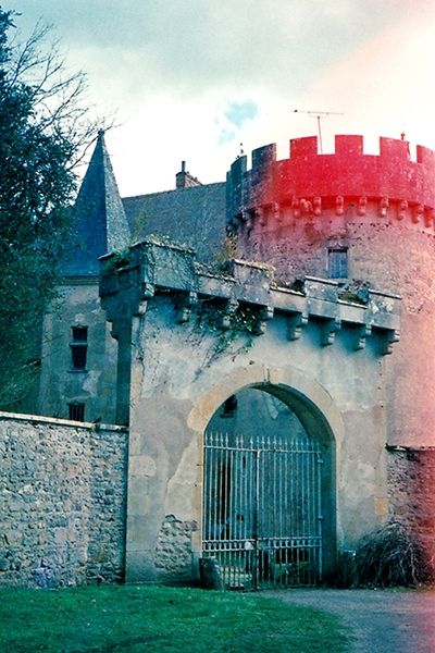 Une grande entrée de château en pierre avec une grille. Derrière on voit un bout du château avec une grosse tour ronde et une carrée.