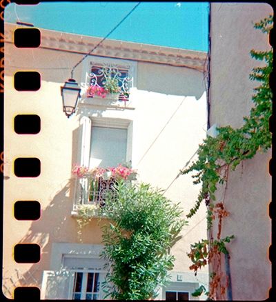 Au croisement de deux rues, une façade avec une fenêtre et un garde corps en fer forgé après lequel sont accroché des jardinières et au dessus une toute petite fenêtre, elle aussi barrée de fer forgé ouvragé.