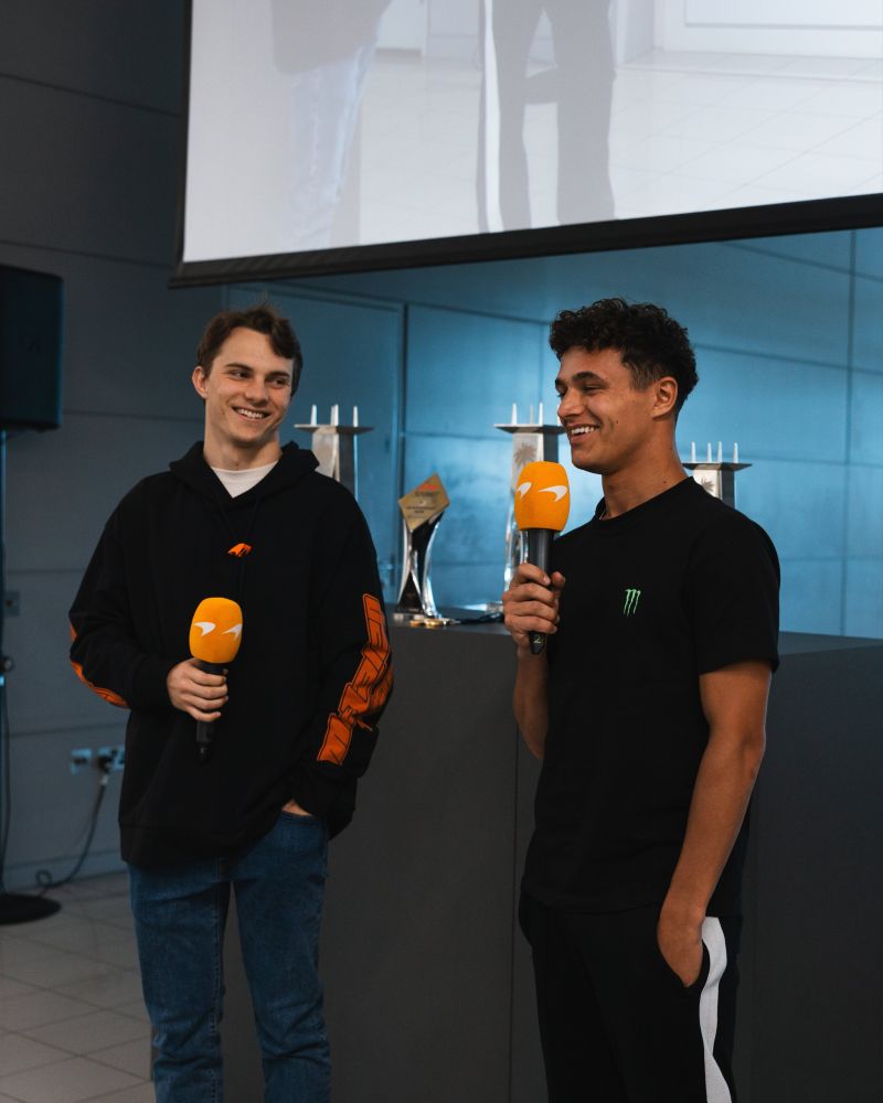 oscar piastri stands on the left in a black hoodie and blue jeans, looking at lando norris on the right, who is wearing a black outfit. they're both holding orange microphones. behind them are the trophies from the miami grand prix